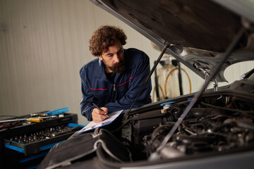 A car mechanic examines an engine while taking notes in a bustling repair garage. Tools are...