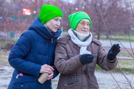 Adult woman walking outdoors with her elderly visually impaired mother in a park. Daughter guiding senior parent during walk. Caregiver assistance for disabled retired woman during autumn