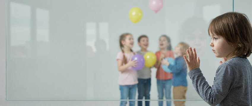 Sad boy behind glass wall labeled Barrier watching happy children play. Social exclusion concept
