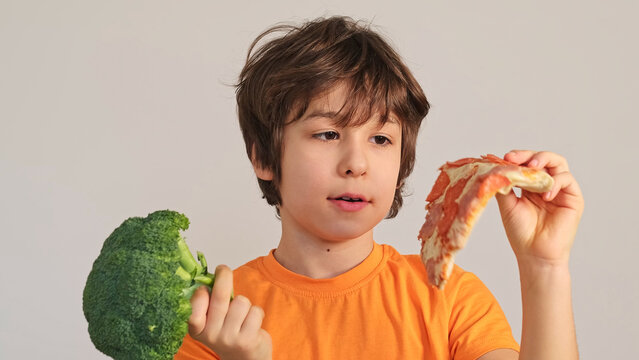 Eye level close up of a cheerful child showing fresh broccoli and a pizza slice against a plain wall. Balanced diet, picky eating, and snack choices matter in everyday family nutrition.