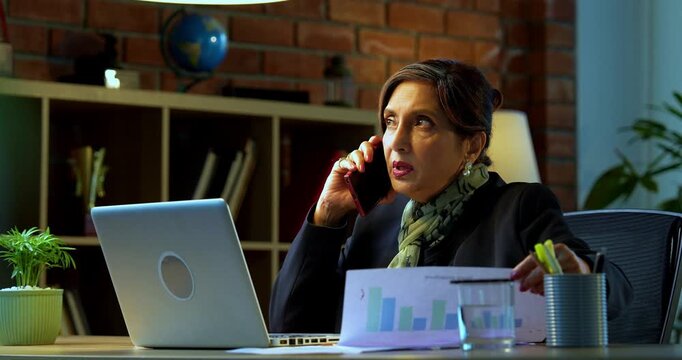 Senior Indian businesswoman in corporate attire attending phone call on smartphone while sitting with laptop and looking at documents in cozy office space experienced female executive at desk