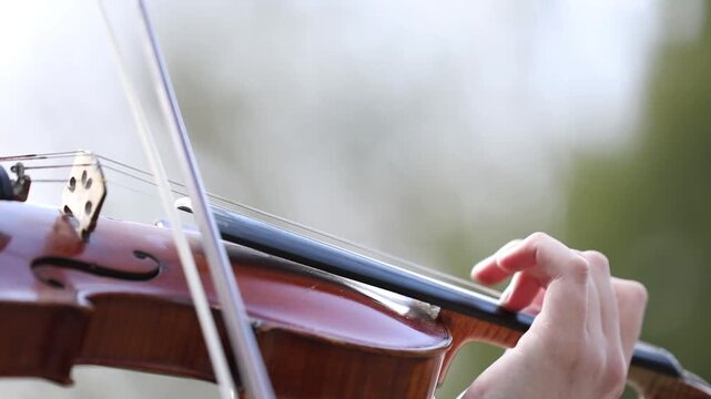 A close up of skilled hands playing a classic wooden violin outdoors. The musician gracefully moves the bow across strings, creating beautiful melodies against a blurred natural background.

