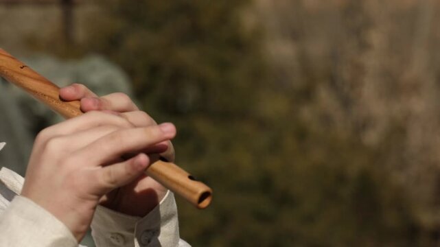 A close-up shot of a musician's hands skillfully playing a traditional wooden flute outdoors. Natural sunlight highlights the instrument and delicate finger movements against a beautifully blurred bac