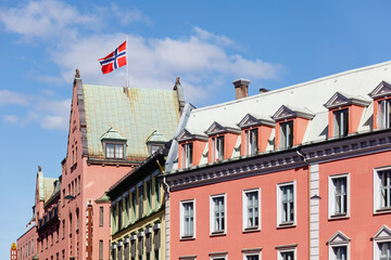 Fototapeta premium Norwegian flag flying above colorful historic buildings in city center- charm of traditional architecture and the patriotic spirit of Constitution Day under a bright blue sky