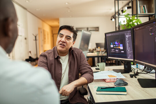 Creative professional mentoring colleague at dual-monitor workstation in modern office