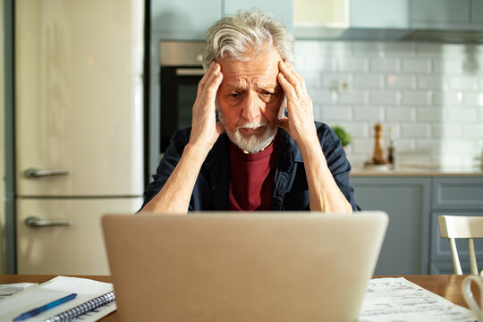Worried senior man checking bills on laptop at kitchen table