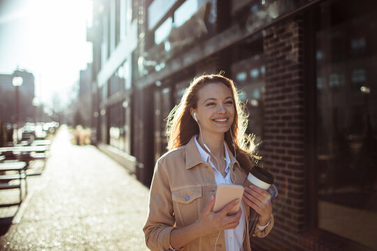 Smiling woman walking with smartphone and coffee on sunny city street