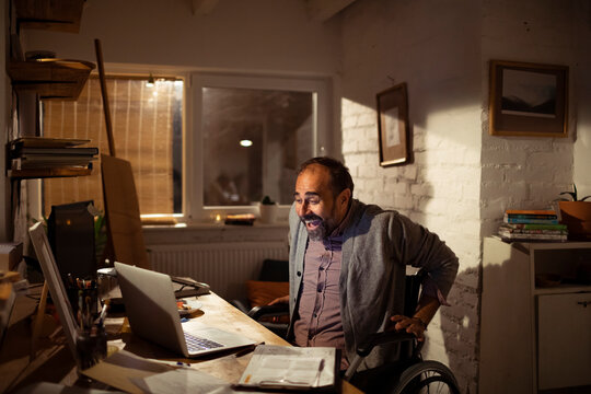 Smiling man in wheelchair video calling in home office at night