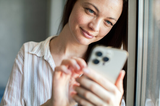 Smiling woman texting on smartphone by window at home