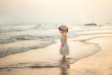 Adorable Asian toddler girl in a white dress exploring the beach shoreline at sunset golden hour. She stands peacefully watching ocean waves during a summer family travel holiday, capturing childhood  © oatawa