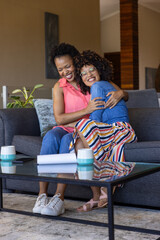 Naklejka premium Diverse female friends sitting on gray sofa in living room, hugging, laughing with mugs