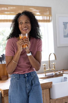 African American woman leaning against wooden kitchen counter holding orange juice by brass faucet