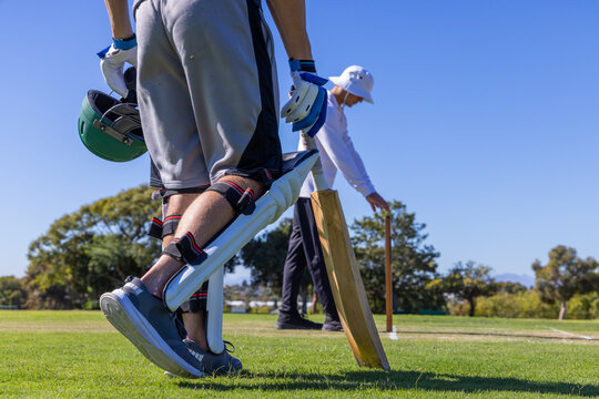 Two male cricketers standing on grass pitch, batter in pads holding helmet and bat near stumps