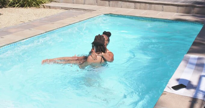 African American couple laughing in yard pool, man lifting woman in bikini splashing to towel phone