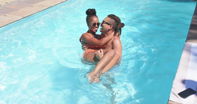 African American couple in backyard pool, man lifting woman celebrating, swimwear phone on deck
