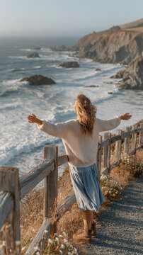 A real vertical 9:16 bright coastal cliff scene, an American white woman standing near a safe wooden overlook fence above the ocean, back facing the camera, arms opened gently as if embracing the sea 