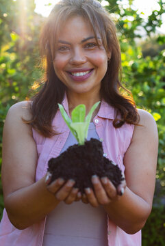 Adult African American woman wearing sleeveless pink shirt holding seedling cupping soil in garden