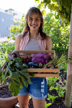 African woman standing in backyard garden holding wooden crate with sweet potatoes, beets, eggplant