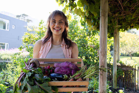 Adult African American woman standing under grapevine canopy holding wooden crate of garden produce