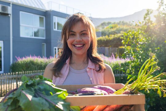 African woman holding wooden crate with carrots and greens in backyard wearing purple tank top