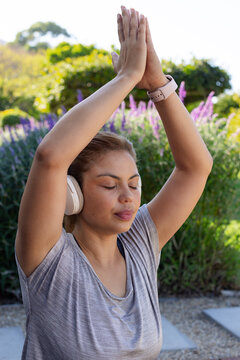 African female standing with arms raised, meditating in garden wearing athletic top and headphones