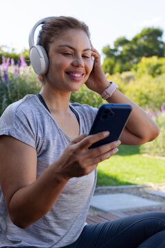 African American woman sitting on step in park adjusting headset, wearing sports top, holding phone