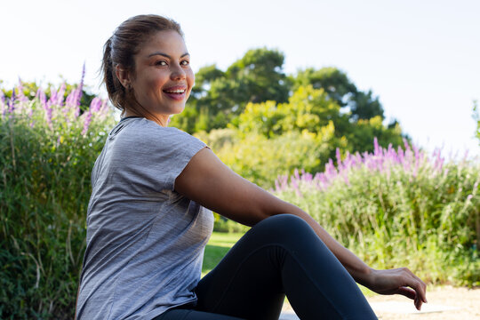 Woman sitting on grassy ground in park wearing grey t-shirt and dark leggings near purple flowers
