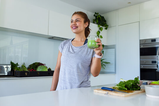 African American woman holding green smoothie at kitchen island, wearing athletic top