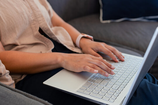 African American woman typing on silver laptop with white keyboard on gray sofa wearing wristwatch