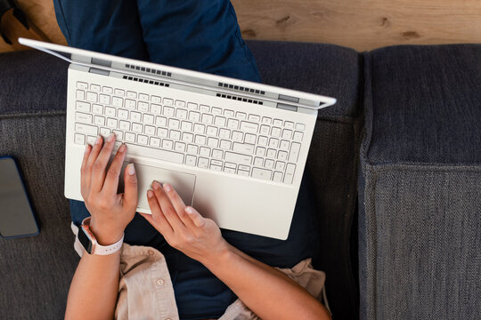 Woman typing on white laptop on dark-gray couch at home wearing beige shirt, smartwatch, smartphone