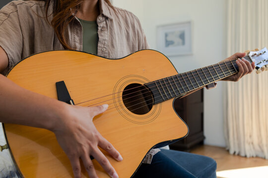 African American woman playing acoustic guitar sitting at home wearing beige shirt and dark jeans