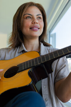 African American woman sitting by window holding acoustic guitar in lap, wearing light button shirt