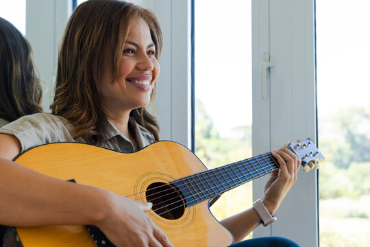 Woman sitting near window in light shirt playing acoustic guitar with mirror, watch