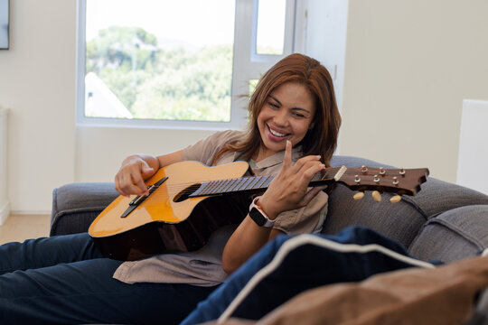 Adult African American woman reclining on gray sofa, playing acoustic guitar in bright living room