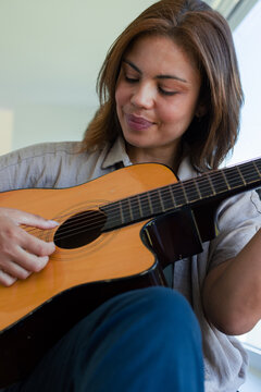 African woman playing acoustic guitar at home, wearing beige button-up shirt and dark blue pants