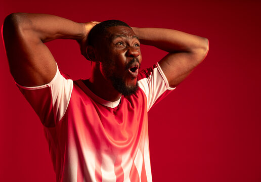 African man standing with hands clasped behind head against red backdrop wearing red-white jersey