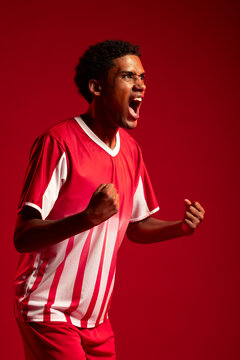Adult African man yelling, clenched fists, in striped jersey, red shorts, studio-lit red backdrop