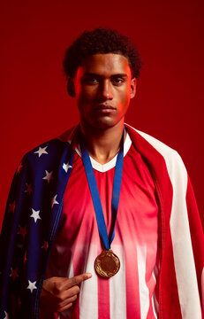 African male athlete pointing to gold medal, wearing American flag, red-and-white jersey in studio