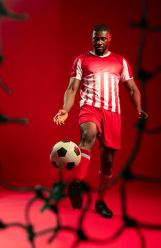 African male soccer player balancing ball in red studio wearing red jersey blurred netting