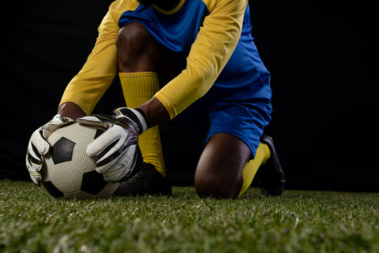 African American male keeper kneeling on studio turf wearing blue kit and white gloves holding ball