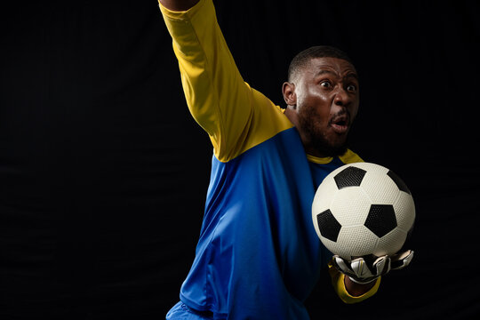 African male goalkeeper in studio wearing gloves, holding soccer ball, raising arm, copy space