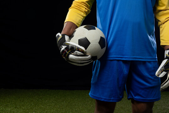 Male keeper holding black-and-white ball at waist, in blue kit, yellow sleeves, gloves on turf
