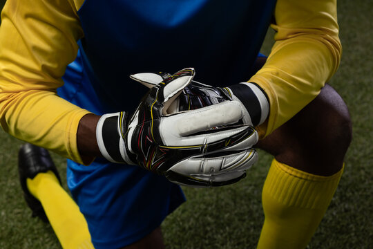 African male goalkeeper kneeling on turf, holding padded white gloves, wearing blue kit