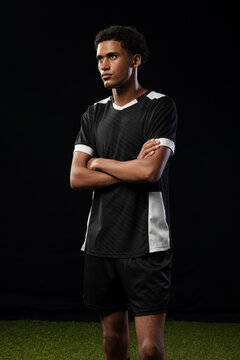 Man standing on turf in studio, wearing black white jersey and shorts against black backdrop