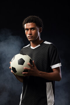 African American male in 20s standing in studio holding soccer ball wearing dark jersey smoky light
