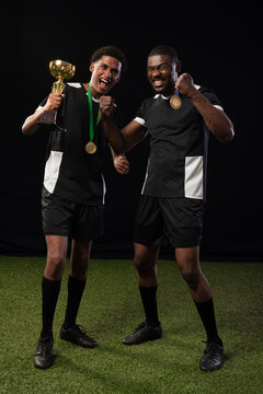 Two male teammates posing on green turf wearing jerseys and gold medals while holding trophy