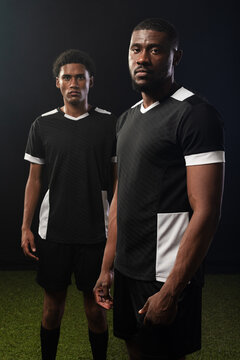 African male teammates standing on turf in studio, wearing black jerseys with white trim and socks