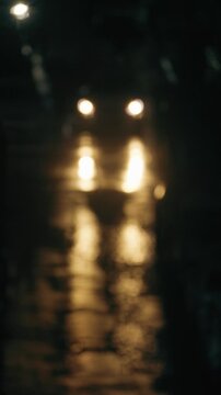 An atmospheric, washed-out look of car headlights at night. Golden bokeh reflections shimmer on the wet, dark asphalt during rain. Dark, cinematic, and mysterious
