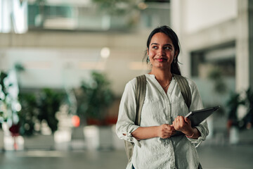 Young indian student smiling and carrying backpack and digital tablet in a modern university campus building