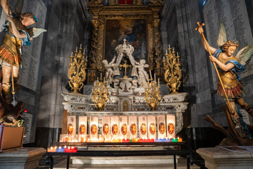 Fototapeta premium Baroque church altar in Menton, France, shows a marble tabernacle with a crown, gilded candlesticks, polychrome archangel statues, candles, and a large painting.